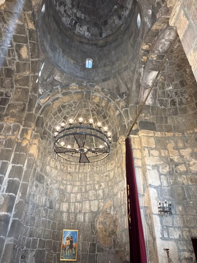 The image displays the interior of a historic stone building, likely a church or monastery, captured with an upward perspective towards its vaulted ceiling and dome. The structure is predominantly built from rough-hewn, irregular grey and brown stone blocks, showcasing an ancient architectural style.
A large, circular metal chandelier dominates the central view, hanging from the dome. It is ornate, featuring numerous illuminated incandescent light bulbs and a central cross-like element fashioned from intricate metalwork. Small, rectangular openings or windows are visible within the curved stone ceiling, allowing some natural light into the space. Several thin metal rods or pipes run across parts of the stone structure.
On the upper right side of the image, a segment of dark red fabric or drapery is visible, draped below a stone ledge. Above it, two decorative metal wall-mounted fixtures are attached to the stone wall, one featuring a grid design and the other resembling a sconce. Below the drapery, a framed painting hangs on the right wall, depicting multiple figures, possibly religious, rendered in light colors against a darker background.
There are no people visible, and no specific activity or event is taking place. The interior is lit by both the prominent artificial light from the chandelier and subtle natural light filtering through the high openings. The overall impression is one of historical significance and solemnity, characteristic of a religious site. The architectural style, particularly the stone work and domed construction, aligns with ancient structures found in Armenia, such as those in Vaghatin. No visible text is present in the media file.
