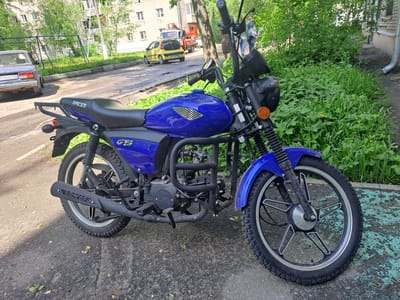A blue Racer moped rests on the pavement near a grassy area in Bel'kovo, Russia. The moped is parked in front of a silver sedan, with a yellow van and an orange cherry picker visible in the background. A building is situated behind the vehicles. There are a few people blurred in the background.