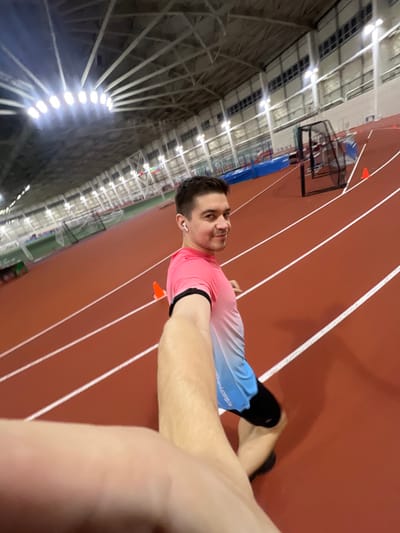 A man takes a selfie at an indoor track. He wears a pink and blue gradient long-sleeve shirt, black running shorts, and white wireless earbuds. A black fitness tracker is visible on his arm. The track is red with white lane markers. An indoor sports complex with a metal frame and bright overhead lights is in the background. There is a basketball hoop near the track. Orange cones are placed on the track.