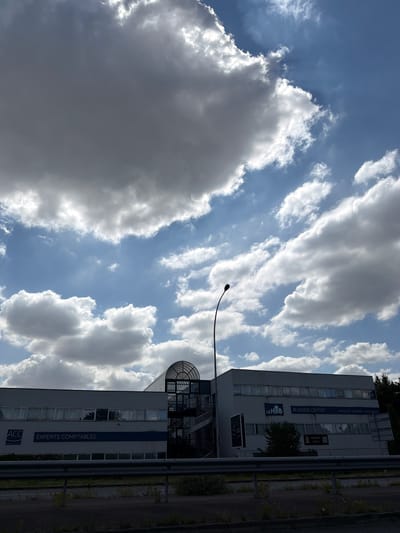 The image depicts a modern office building in Évry-Courcouronnes, France, with a backdrop of a dynamic sky filled with both dark and bright clouds. The building, which appears to be a business center, has large windows and prominent signage including "ACC EXPERTS COMPTABLES" and "LE HUB BUSINESS CENTER." An external spiral staircase is visible on the left side of the building, leading up to a curved glass structure. In the foreground, a metal barrier separates the road from the building's property. A street lamp with a distinctive curved design is positioned to the left of the building, silhouetted against the sky. The overall scene is captured from a slightly elevated perspective, possibly from a vehicle on the adjacent road.