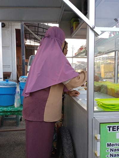 A woman wearing a pink hijab and a maroon and tan outfit is standing behind a metal food cart, potentially preparing food for a customer. The cart contains various green containers filled with food items. Stacks of green plates are visible on top of the counter. Behind the woman, there is a tire leaning against the cart. The location appears to be outdoors, possibly a street in Pilang, Indonesia, as suggested by the urban environment visible in the background. Other elements such as blue plastic water tanks and buildings are also visible in the scene.