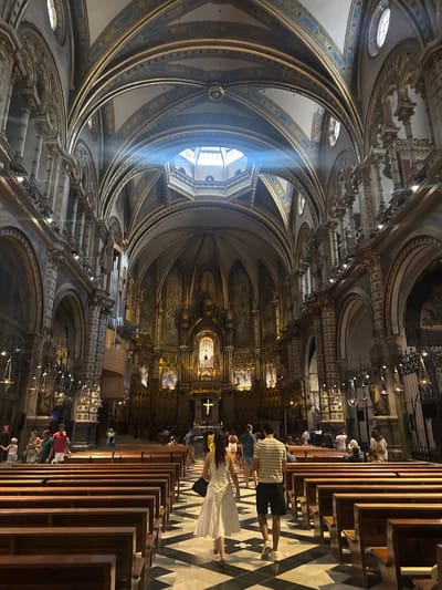 The image shows the interior of a grand cathedral, likely the Benedictine Monastery of Montserrat, located in the municipality of Collbató, Spain. The perspective is from the nave, looking towards the altar. Sunlight streams through a high opening, illuminating dust motes in the air and casting a soft glow on the ornate ceiling.
The architecture is characterized by soaring vaulted ceilings with intricate golden detailing and blue accents. Large arched windows punctuate the upper walls. The walls themselves are decorated with religious imagery and elaborate carvings. A large pipe organ is visible on the left side of the nave.
In the foreground and mid-ground, rows of dark wooden pews are arranged on a patterned tiled floor, alternating black and white squares. Several individuals, appearing to be tourists, are scattered throughout the nave. A couple in the mid-right foreground walks away from the camera towards the altar; the woman is wearing a white dress, and the man is in a striped shirt and shorts. Other people are seen standing or sitting in the pews, or walking along the aisles. The overall atmosphere is one of reverence and awe, with the architecture and light contributing to a majestic ambiance.