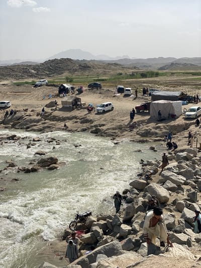 The image depicts a riverine landscape with a congregation of people, vehicles, and tents. The river, characterized by its rocky bed and turbulent flow, forms a prominent part of the foreground. People are situated both along the riverbank, climbing on rocks, and further inland, near makeshift encampments.

A mix of vehicles, including white cars and a red truck, are parked on the terrain away from the river's edge. Tents of various sizes are erected nearby, suggesting a temporary settlement or gathering. The background shows a landscape featuring elevated terrain and a somewhat hazy sky.

The presence of people engaging in various activities near the river and the setup of tents indicates this is a populated area. The location is identified as Kim, Afghanistan.