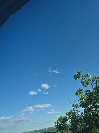 The image depicts a bright, clear blue sky with scattered white clouds. A portion of a dark, textured object, possibly a roof or overhang, occupies the upper left corner of the frame. In the lower right section of the image, a lush green tree branch extends into the sky, its leaves vibrant and full. In the far distance, a hazy, verdant landscape is visible, suggesting a rural or semi-rural setting. A faint crescent moon can be seen in the upper portion of the sky. The overall impression is one of a sunny day in Merenii Noi, Moldova.