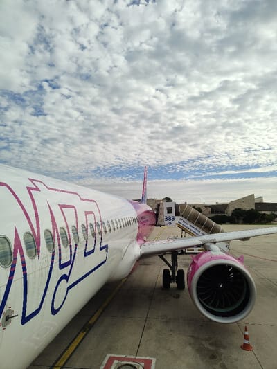 A Wizz Air Airbus aircraft is parked on an airport tarmac under a partly cloudy sky. The aircraft's fuselage is white with large purple "WIZZ" lettering, and its tail fin and engine nacelles are also purple. Visible features include multiple circular passenger windows along the fuselage and the forward-left engine with its internal fan blades. A mobile passenger jet bridge, labeled "383" and bearing text in Hebrew "נמל התעופה בן גוריון" (Ben Gurion Airport) and partial English "GEA GURION INTERNA", is connected to the aircraft's forward door. In the foreground, a red and white traffic cone is placed near the aircraft's engine, and yellow tarmac markings are visible on the concrete surface. In the background, modern airport terminal buildings with stone or concrete facades are discernable. The sky is characterized by numerous patchy white clouds against a blue background. No individuals or specific actions are discernible. The scene is located at Ben Gurion Airport, Israel, which is situated near Yagel.
