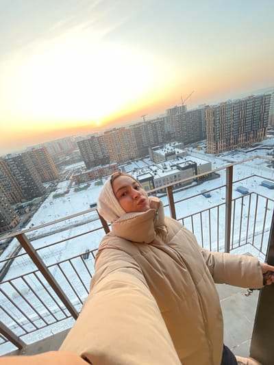 A female individual, approximately 20-35 years old, is depicted in a selfie taken on an elevated outdoor balcony in Elektrougli, Russia. She is wearing a light beige hooded puffer jacket, with a beige fabric element covering part of her head and neck. Her lips are pursed, and her eyes are directed towards the camera. Her right hand is extended, presumably holding the camera device, while her left arm rests against a dark brown metal balcony railing.

The background presents an expansive cityscape under winter conditions, with all visible ground surfaces covered in snow. Numerous multi-story residential buildings of varying modern architectural styles, predominantly in brown, grey, and white tones, populate the urban landscape. Several construction cranes are visible in the distance among the buildings, indicating ongoing development. Lower institutional-type buildings are also scattered throughout the scene. The sky occupies the upper frame, transitioning from a bright, intense golden-orange hue on the left horizon, where the sun is visible as a distinct disc, to a lighter blue-grey overhead. The low position of the sun suggests either sunrise or sunset.