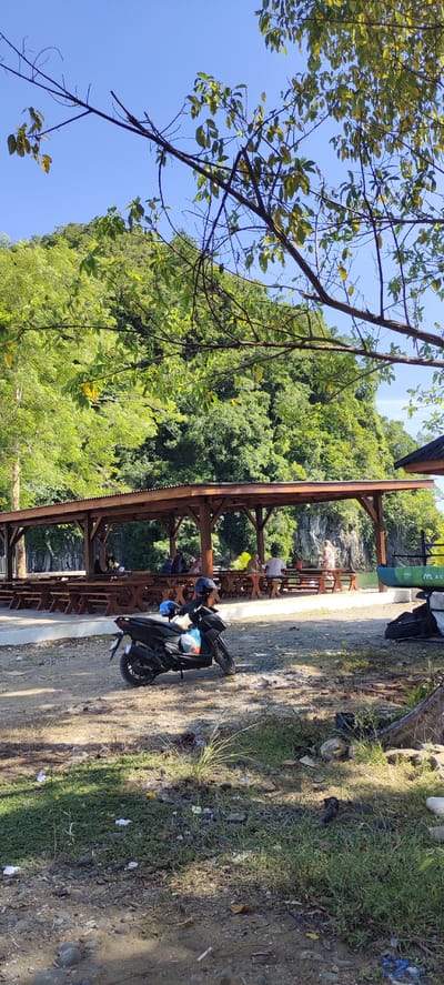 A sunlit, outdoor scene captures a recreational area near a body of water, likely in or around Lamseunia, Indonesia. In the foreground, a dark-colored scooter is parked on a gravel and dirt path. The scooter has a blue helmet secured to its handlebars and a plastic bag hanging from it. Behind the scooter, a shaded open-air pavilion with a wooden frame and a corrugated metal roof extends across the middle ground. Several long picnic tables with benches are arranged under the pavilion, with a few people seated at them. The people appear to be in casual attire and are either conversing or resting.

Beyond the pavilion, a calm body of water is visible, with a lush, green, tree-covered hillside rising dramatically behind it. The trees are dense, suggesting a tropical or subtropical environment. A portion of a green kayak with white lettering is visible on the right side of the image near the water's edge. The sky is clear and bright blue. The overall impression is a relaxed atmosphere, possibly a park or a picnic spot popular with locals.