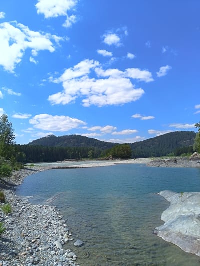 The image depicts a scenic natural landscape featuring a wide, calm river with clear, shallow water in the foreground. The riverbed is visible, lined with numerous smooth, gray and brown pebbles. Large, flat gray rocks emerge from the water's edge on the right side of the frame. The riverbanks are covered with a dense layer of pebbles, transitioning into green vegetation and trees further back. In the middle ground, the river flows past a rocky island and a line of trees, with a gravel bar extending into the water. The background is dominated by rolling, tree-covered hills, their slopes densely packed with dark green coniferous trees. Above the hills, a vibrant blue sky is dotted with scattered white, fluffy cumulus clouds. The lighting suggests it is daytime, likely midday, with the sun illuminating the scene. There are no people or animals visible. The location is identified as Nizhnekuyanchinskoye, Russia.