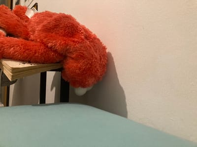 A large, bright orange, plush toy, possibly a stuffed animal resembling Elmo, is lying face down on a small table. Its head and upper body are hanging off the edge of the table. The table is made of wood and has a black metal base. It is partially obscured by a shadow cast on the wall. The wall is white and textured. The toy’s shadow is cast on the wall. A soft, light blue, fabric surface, possibly a cushion or blanket, is visible at the bottom of the image. The toy is in the foreground, the table is in the middle ground, and the wall is in the background. The location is a residential building in Kokhav Ya'ir Tzur Yigal, Israel, at coordinates 32.209, 34.989.