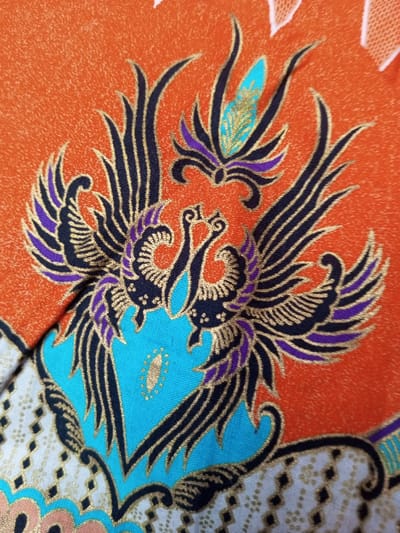 The image displays a segment of vibrant Batik fabric. A pair of stylized phoenixes, depicted in black with gold accents and purple highlights, dominates the composition. They are positioned atop a turquoise base. The backdrop features a textured orange field, broken by white sections decorated with geometric patterns. Another turquoise element with gold accents is included.