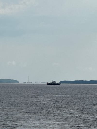 The image depicts a wide, gray body of water under a cloudy sky. On the water, a dark-colored vessel, appearing to be a tugboat or a small freighter, is visible in the middle distance. To the left of the vessel, a thin, tall tower structure, possibly a radio mast or a distant lighthouse, rises from a distant landmass. The landmasses on the left and right appear as dark, tree-covered shapes on the horizon. The water is textured with ripples and waves. The overall atmosphere is muted and overcast.