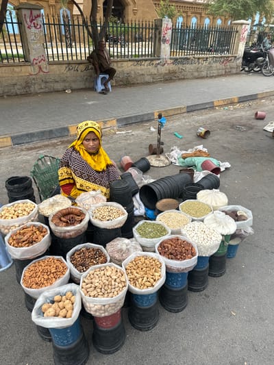 A woman in a yellow and black headscarf and traditional clothing is sitting on a mat amidst a collection of bags filled with various nuts and dried fruits. The bags are stacked in rows, some resting on large black cylindrical containers, others directly on the ground. The contents of the bags include what appear to be pistachios, almonds, walnuts, dried figs, and other unidentifiable items. The scene is set on a paved street, with a metal fence and buildings in the background. A man is visible sitting on a small blue stool in the background, to the left of the woman. Several motorcycles are parked near the fence. The ground is dirty and littered with some debris, including plastic bags and discarded containers. The lighting suggests it is daytime, likely morning or afternoon, with diffuse light indicating an overcast or partially cloudy sky. The presence of local architecture and the clothing of the individuals suggest a location in Karachi Division, Pakistan. The woman's gaze is directed towards the viewer, and her expression is neutral. The overall scene depicts a street vendor or a stall at a market.
