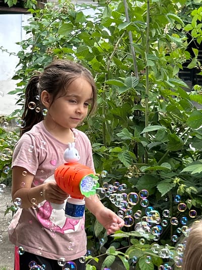 A young girl is holding a bubble gun in the shape of a carrot and is blowing bubbles. She is wearing a pink t-shirt with a cartoon character on it. The background is filled with lush green foliage, suggesting an outdoor setting. The lighting indicates it is daytime, and the presence of leaves suggests a warm season. There are many iridescent bubbles floating around the girl. The image appears to be taken in a residential area in Prokhladny, Russia, possibly a backyard or garden. The girl's expression is a mix of concentration and enjoyment as she engages with the bubbles.