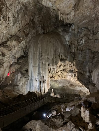 The image depicts a cave interior, likely a tourist attraction, with prominent rock formations, stalactites, and stalagmites. A metal staircase ascends along one of the cave walls, possibly providing access for visitors. A small red light is visible at the bottom of the frame, potentially marking a path or hazard. A brighter white light illuminates an area higher up, possibly a natural opening or artificial lighting fixture. The environment is characterized by the textures and colors of natural stone, with varying shades of gray and brown. The location is purportedly in Inkiti, Abkhazia.