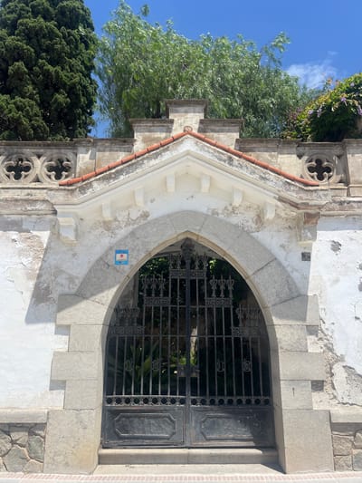 This vertical image depicts a weathered architectural facade, likely a building in Sant Pol De Mar, Spain, under bright daylight. A prominent feature is a pointed stone archway made of light-colored blocks, framing a dark, ornate wrought-iron grille. The grille features intricate scrollwork and circular designs, allowing glimpses of green foliage behind it.
The surrounding wall is primarily white, exhibiting signs of age with peeling paint and discolored patches. To the left of the arch, a series of decorative white stepped elements descend, topped by terracotta-colored tiles. Further left, a stone column incorporates quatrefoil cutouts, suggesting a Gothic or neo-Gothic influence. The base of the wall on the right side includes a section of rough-hewn stone blocks.
Above the structure, a clear blue sky with a few wispy white clouds is visible, along with abundant green trees and shrubs, some bearing small purple flowers. The bright sunshine indicates it is daytime, likely mid-morning or afternoon, as evidenced by the distinct shadows.
A small, square blue and white tiled plaque is affixed to the wall beneath the apex of the arch. While difficult to read entirely, a red sun symbol is visible on the plaque, and the text "SANT POL DE MAR" is partially discernible at the bottom, confirming the location. The scene is devoid of human activity, conveying a sense of historical tranquility and natural beauty.