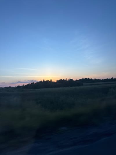 The image depicts a landscape scene during twilight, likely near Orlivka, Ukraine. The sky is a gradient of deep blue at the top, transitioning to lighter blues, pinks, and oranges near the horizon, indicating either sunrise or sunset. A thin line of clouds is visible horizontally across the sky. Below the sky, a dark silhouette of a treeline forms a distinct horizon. In the foreground, a field is visible, appearing dark and somewhat blurred, suggesting motion or low light conditions. The bottom portion of the image is very dark, showing a blurred texture that could be a road or another dark surface. There are no people, animals, or specific man-made structures clearly identifiable in the frame. The overall impression is one of a quiet, natural landscape at dawn or dusk.