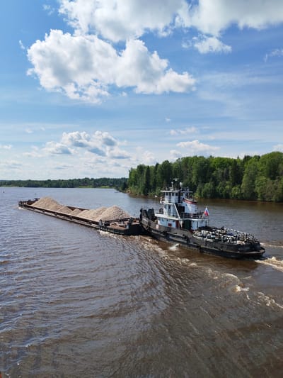 A tugboat named "ШЛЕЗОВСКОЙ-8" is pushing a barge loaded with wood chips along a brown river. The scene is likely located near Kurikaevo, Russia. The water is choppy from the boat's wake. Green trees line the riverbanks in the background, and a blue sky with fluffy white clouds is above.