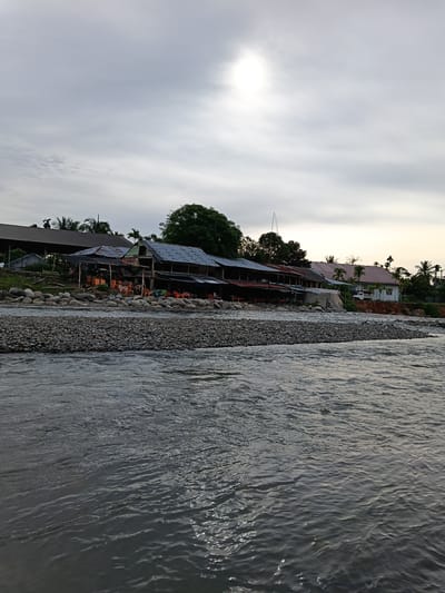The image depicts a wide, grey river in the foreground, with ripples and reflections on its surface. The riverbank is lined with numerous grey and brown stones. Beyond the riverbank, a row of buildings with corrugated metal roofs is visible. Some of these buildings appear to be open-air cafes or restaurants, with tables and chairs set up outside. Trees, including palm trees, are scattered among the buildings and behind them. The sky is overcast, with a bright, diffused sun appearing in the upper center of the frame. The scene suggests a riverside settlement, possibly in a tropical or subtropical region. The location is identified as Bateiiliek, Indonesia.