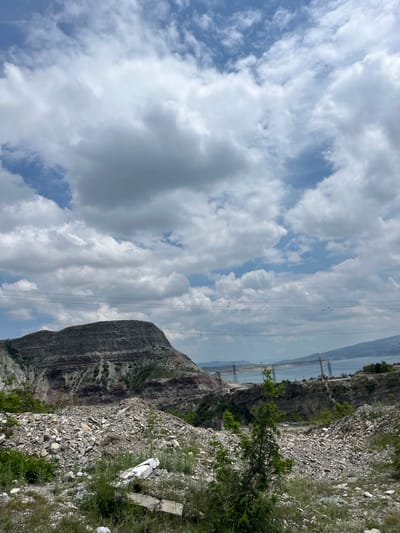 The image depicts a mountainous landscape under a partly cloudy sky in Chirkey, Russia. A large, striated rock formation dominates the foreground, with power lines stretching across the frame towards what appears to be a lake or reservoir in the background. The foreground also includes rocky terrain with sparse vegetation. In the distance, a structure resembling a bridge or dam can be discerned. No people are visible in the image.