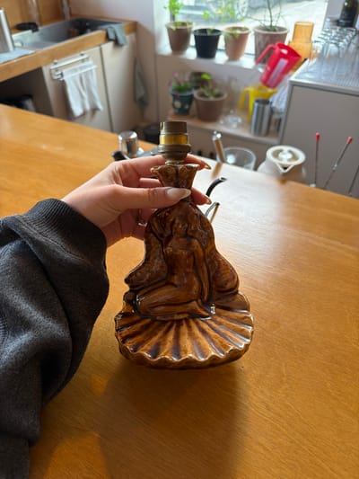 A person is holding a brown ceramic lamp base in their hand, which is adorned with a high-relief design of a reclining nude figure and a stylized shell pattern on the broader end. The lamp base features a threaded metal fitting at the top, presumably for attaching a lampshade. The person's hand has manicured nails and a ring is visible on one finger. The individual is wearing a dark grey hooded sweater. The background features a light brown wooden table surface. In the distance, there is a kitchen area with visible kitchenware, plants, and light-colored surfaces. This image does not provide any evidence to verify that it was taken in Toseong-myeon, South Korea.