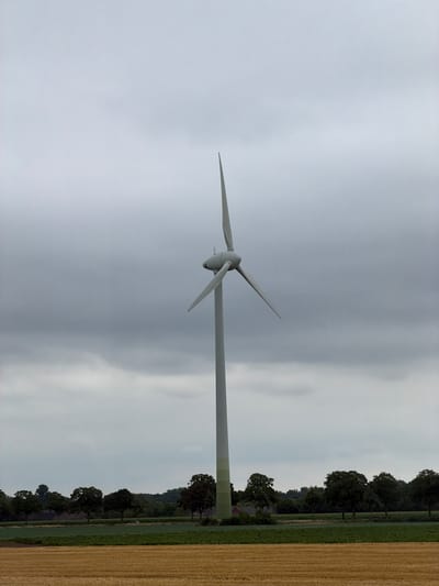 A single white wind turbine stands vertically in the center of the frame, its three blades angled upwards against a cloudy, overcast sky. The turbine's tower has a light green base and a white upper section. In the background, a line of dark green trees forms a horizon, behind which a faint outline of rolling hills is visible. In the foreground, a strip of green field is present, separated from the viewer by a field of dry, golden straw. The scene is located in the rural area of Kerken, Germany.