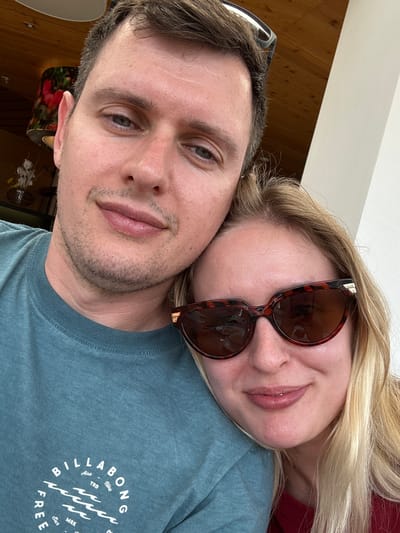 A close-up selfie shows a man and a woman. The man, on the left, has short brown hair, blue eyes, and is wearing a teal t-shirt with a Billabong logo. He is looking at the camera. The woman, on the right, has long blonde hair and is wearing sunglasses with tortoiseshell frames. She is leaning her head on the man's shoulder and smiling. The background shows a wooden ceiling and a white wall, suggesting an indoor setting. The scene is likely within the city of Heiligenblut am Großglockner, Austria, given the prompt.