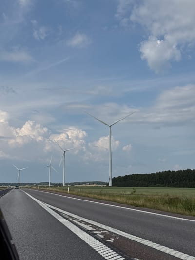 The image depicts a highway scene with multiple wind turbines in the background, located near the city of Åbytorp, Sweden. The highway is paved with asphalt and has white lane markings. The wind turbines are white and tall, with three blades each, and are positioned on a grassy field behind a treeline. The sky is blue with scattered white clouds. The overall impression is of a rural or semi-rural landscape under a daytime sky.