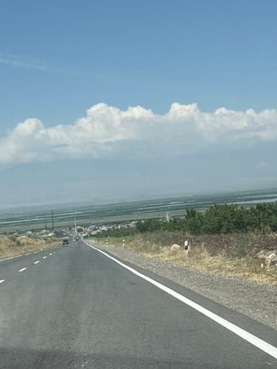 The image captures a view of a road in Paruyr Sevak, Armenia. The road is paved and has a white line marking its edge and a dashed white line dividing the lanes. There are a few cars driving on the road, and some utility poles are visible in the distance. The landscape features dry grass and bushes on the sides of the road, with a town or village in the middle ground. In the background, there are expansive fields, a body of water, and distant mountains, all under a bright blue sky with scattered white clouds.