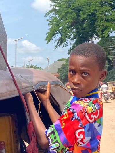 In Igbara, Nigeria, a young Black boy with short hair and dark eyes, wearing a vibrant, multi-colored shirt featuring the "TikTok" logo, is positioned in the foreground, looking directly at the camera. He is leaning against a weathered, brown vehicle, possibly a tricycle or tuk-tuk, with his hands visible on its surface. The boy's shirt is adorned with various colorful shapes and the text "TikTok" prominently displayed in a rainbow of colors. Behind him, the environment suggests a daytime outdoor setting with a dusty ground, trees with lush green foliage, and a bright blue sky with some clouds. Several streetlights are visible in the background, along with a building. Further in the distance, people and motorcycles can be seen, indicating activity on a street or marketplace. The lighting suggests it is a sunny day.