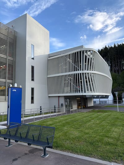 This image captures a modern architectural scene in Troisvierges, Luxembourg, under a clear blue sky with scattered white clouds, indicating daytime and good weather.

The primary subject is a contemporary building featuring a distinctive light-colored, curved facade composed of horizontal slats, possibly serving as a ventilation or sun-shading element. Below this prominent feature, the building has a more solid, rectangular section with several small, horizontal windows. The structure appears clean and well-maintained.

Surrounding the building, a neatly manicured green lawn with visible mowing stripes extends alongside an asphalt pathway or road. In the background, a dense cluster of dark coniferous trees provides a natural backdrop.

In the foreground, a dark-colored metal bench with a grid-patterned seat and backrest is fixed to the asphalt, positioned next to the grassy area. Adjacent to the bench, a bright blue, rectangular, box-like object stands prominently. This object displays the white capital letters "CFL" on its side, identifying it with Chemins de Fer Luxembourgeois, Luxembourg's national railway company, consistent with Troisvierges being a significant railway location. Metal railings are visible along the path leading towards the building's entrance.

The scene is devoid of people or specific activities, presenting a calm, orderly, and modern outdoor environment.