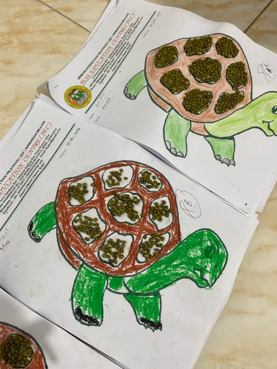 The image shows two crayon drawings of turtles on white paper. The turtles have brown shells filled with green mung beans. The drawings are placed on top of some papers that have text in Indonesian, indicating they are from "JESS EDUCATION CENTRE (JEC)" located at "Jl. Pierre Tendean Kel. Tanjung Gading Kec. Pasir Puyu, Inhu". This location is in the city of Tanjung Gading, Indonesia. The drawings appear to be art projects completed by children. The lighting suggests an indoor setting.