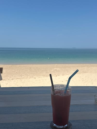 A tall, transparent glass containing a red-pink liquid with external condensation rests on a dark coaster atop a multi-planked wooden surface in the foreground. Two straws, one dark and straight, one light blue and bent, protrude from the liquid. The wooden surface consists of alternating light blue and light gray horizontal planks. A small, clear glass ashtray is partially visible to the right. The midground presents an expansive, unoccupied light-tan sandy beach. The background features the Andaman Sea, exhibiting a gradient from light turquoise near the shore to a darker blue further out. A single white buoy floats in the water towards the middle-left. A small, dark landmass is discernible on the distant horizon, towards the right. The upper sky is clear and uniformly blue. This scene is situated in Phra Ae, Thailand.