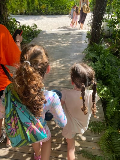The image shows an outdoor scene with a wooden walkway. Two young girls are in the foreground, one with braids and wearing a light-colored shirt, and the other with curly hair tied in a ponytail, wearing a colorful shirt and carrying a reusable shopping bag with a floral pattern. An adult wearing an orange shirt is holding a phone. In the background, other people can be seen walking on the path. Lush greenery surrounds the walkway. The location is likely Marina Maroma, Mexico, based on the user's prompt.
