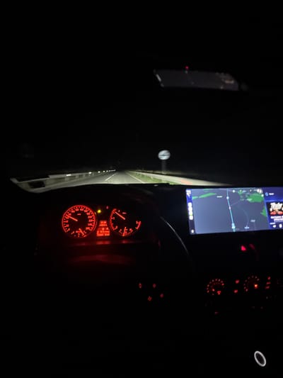 This is a photograph taken from inside a moving car at night. The dashboard is visible, displaying illuminated red gauges, including a speedometer and other indicators. A screen displaying a map is visible in the upper left portion of the photograph. The road is visible through the windshield, illuminated by the headlights of the car. The OCR data indicates that the time is 23:34, the outside temperature is 19.5 degrees, and that the car is in D4 gear. The location is likely Mogila, Bulgaria, due to the text "Tur" (likely "Turn") being displayed on the navigation screen, though this cannot be definitively confirmed.