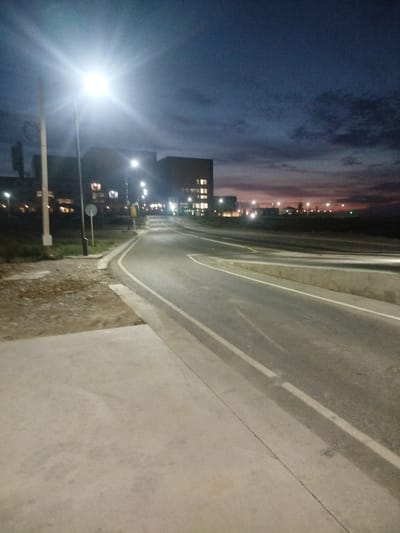 The image depicts an outdoor scene at dusk, characterized by artificial lighting and a twilight sky. In the foreground, a concrete sidewalk runs alongside a paved road. The road curves to the right, with white lane markings visible. To the left of the road, there is a patch of rough ground with gravel and some sparse vegetation, bordered by a concrete curb. Further back, a lamppost with a bright light fixture is prominent on the left side. Beyond the road, several multi-story buildings with illuminated windows are visible. The sky above is a deep blue with wisps of clouds, transitioning to a lighter, reddish hue near the horizon, suggesting the end of daylight. The overall environment appears to be an urban or semi-urban area, likely a road or highway with adjacent structures. The specific location, Түндүк айланып өтүүчү жол 84 га, Kyrgyzstan, indicates a northern bypass road.