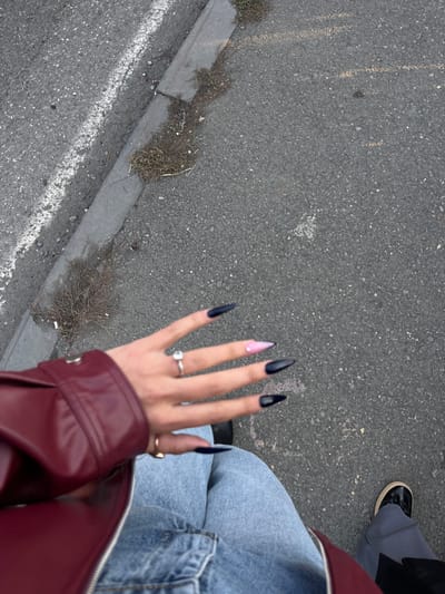 A high-angle, downward-looking shot captures a person's left hand, partially visible torso, and legs, alongside the lower leg and foot of a second individual. The primary subject's left hand features long, pointed (stiletto) nails; four are solid black, while the middle finger nail is pink with a lighter tip. A silver-toned ring with a clear stone is on the ring finger, and a gold-toned ring on the thumb. The subject wears a dark red leather-like jacket with a silver snap button and blue denim jeans. To the bottom right, a second individual's right foot, wearing a black shoe with a thick white sole, and a grey textured pant leg are visible. The ground consists of dark grey asphalt pavement containing scattered light-colored aggregate, marked by a faded white line running diagonally. A dark grey concrete curb, partially overgrown with dry, sparse weeds and grass, borders the asphalt. No specific landmarks are visible to determine the location within the city of Lanchkhuti, Georgia.