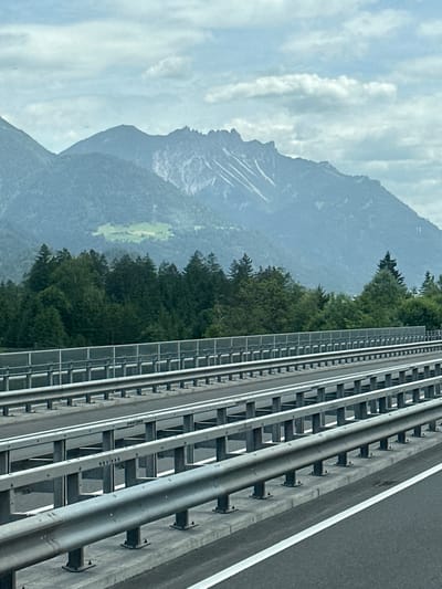 The image depicts a highway with metal guardrails, indicating a controlled-access road. In the background, a dense forest of evergreen trees forms a dark green band. Beyond the forest, imposing mountains rise, characterized by rugged peaks and some patches of snow or lighter rock. The sky is overcast with scattered clouds. The perspective suggests the photo was taken from a moving vehicle, likely a car, due to the slightly blurred appearance and the focus on the road infrastructure. The scene appears to be in a mountainous region, consistent with the geography of Nenzing, Austria, which is situated in the Rhine Valley with access to alpine landscapes. There are no people or specific urban elements visible in the image, focusing instead on the natural environment and the infrastructure of the highway.