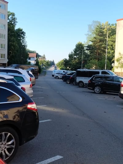 The image captures a street scene in Písek, Czechia, under clear blue skies during daylight hours, likely late afternoon due to the warm, low-angle sunlight casting long shadows. The main subjects are a row of parked cars on an asphalt road, demarcated by white lines. The cars are varied, including a black van with white lettering that reads "KLIMATIZACE - TEPELNÁ ČERPADLA" (Air Conditioning - Heat Pumps), suggesting a service vehicle. Other vehicles include a black SUV in the foreground and several other cars and vans further down the street.

The setting features residential buildings with a light-colored facade and a reddish-brown tiled roof visible on the left side of the image. Lush green trees line the street and the area around the buildings, indicating a well-treed neighborhood. The presence of streetlights and what appear to be utility poles suggests a typical urban or suburban environment. The overall mood is tranquil and ordinary, depicting a quiet moment in a residential area. No people are visible in the image, and the focus is entirely on the parked vehicles and the surrounding environment.