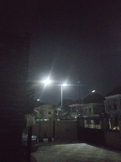 A low-light, outdoor scene depicts a street at night in Rumuomasi, Nigeria. The primary light source is a bright, central streetlamp, casting a strong, white glare and lens flare. To the left, a dark, brick-like structure dominates the foreground, with a partial view of a white object hanging on its side. In the midground, the street continues, lined by a brick wall and fence. Beyond the wall, houses with multiple windows are visible, some illuminated from within. Another streetlamp, less bright than the central one, is positioned further down the street to the right. A small, dark-colored vehicle is partially visible in the lower left of the image, parked near the brick structure. The ground appears to be paved with cobblestones. The sky is dark and overcast, contributing to the overall dimness of the scene.