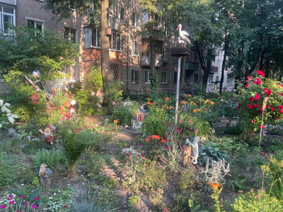 The image depicts a small, overgrown garden in front of a multi-story brick apartment building, likely in the city of Chyornoe, Russia. The garden is filled with a variety of flowers, including red roses, orange lilies, and pink and white smaller blooms. Several garden gnomes and animal figurines are scattered throughout the garden, including two storks on a nest atop a pole, two rabbits on a stump, a gnome holding a sign that reads "ПРИВЕТ" (Hello), and a cat figurine. A large tree stands to the right of the building, casting shadows over the scene. The building itself appears to be made of light-colored brick with multiple windows, some with balconies. The overall impression is one of a well-loved, albeit somewhat wild, urban garden.