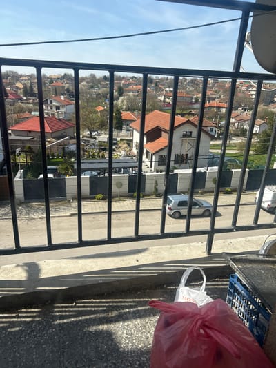 The image shows a view from a balcony in Valchi Dol, Bulgaria, looking out over a residential area. The foreground is dominated by the metal railings of the balcony. To the right of the railing is a plastic shopping bag, with contents in a red bag inside, and a blue plastic crate. The background reveals a mix of houses with red-tiled roofs, trees with sparse foliage, and streets with parked cars. The sky is light blue and clear. The scene appears to be daytime and sunny.