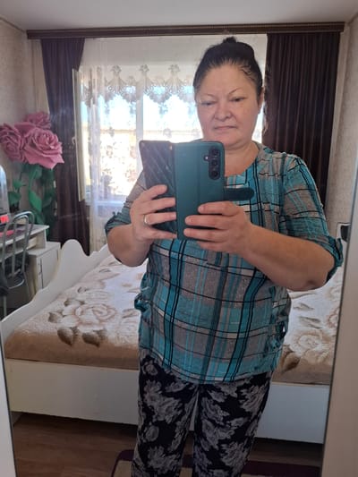 A woman is taking a mirror selfie in what appears to be a bedroom. She is wearing a blue and grey plaid shirt and black pants with a floral pattern. She is holding a dark green phone case in her right hand, which is positioned in front of her chest. A ring is visible on her left ring finger. The background shows a bed with a floral patterned blanket, a window with sheer curtains, and a dark curtain. There are also some pink artificial roses visible on a surface in the foreground, reflected in the mirror. The overall lighting suggests it might be daytime. The reflection in the mirror also reveals a small dresser or drawer unit. The scene is likely indoors, within a residential setting.