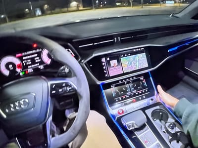 This image shows the interior of a modern Audi vehicle, likely at night, as indicated by the illuminated dashboard and ambient lighting. The dashboard features a digital instrument cluster with various readouts, a central infotainment screen displaying navigation and media, and a touch-sensitive climate control panel. The steering wheel is partially visible, with controls on its spokes. A person's hand is visible on the center console, near the cup holders and gear selector. The surrounding environment appears to be an outdoor parking lot or street, with blurred lights and a dark, possibly paved surface. The location of Bezden, Bulgaria is not identifiable from the visual information in the image itself.