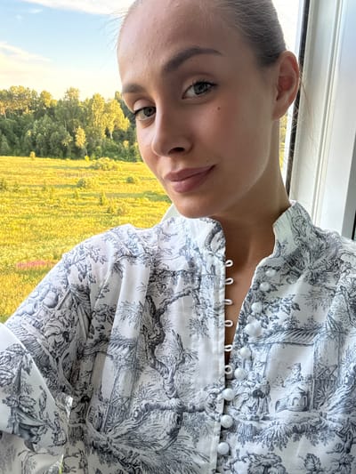 A young woman with light brown hair pulled back in a bun is looking at the camera with a slight smile. She is wearing a white shirt with a black toile print of rural scenes. The background shows a sunlit yellow field and a line of trees under a blue sky. The image appears to be taken outdoors during the day. The location of Шолохово, Russia is not discernible from the image.