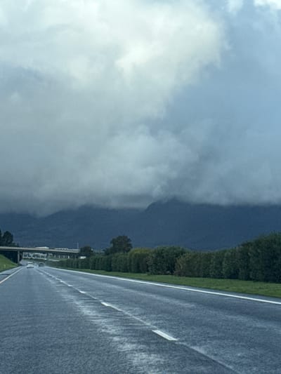 The image depicts a view from a vehicle traveling on a wet asphalt road in Paarl, South Africa. To the right is a dense line of green shrubbery bordering the road. Further away are what appear to be buildings, but they are small and indistinct. The left side of the image is dominated by dark, heavy clouds. The weather is overcast with rain evident on the road surface.