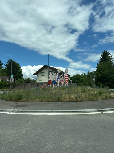 A vibrant outdoor scene, likely captured in Zurzach, Switzerland, depicts a roadside adorned with playful cartoon cutouts of people and a dog. The image, best viewed in landscape orientation, is set under a bright blue sky filled with scattered white cumulus clouds.

In the foreground, a gray asphalt road with a white lane marking and a concrete curb runs along the right side of the frame, bordered by a strip of green grass and weeds to its left. The midground features five flat, cartoon-style cutouts of individuals and a small dog, arranged in a line as if standing or frolicking. These figures appear to be dressed in swim attire, including an adult male in red and white striped shorts, an adult female in a blue swimsuit, two children (one holding a beach ball), and another adult male in red shorts, accompanied by a small brown and white dog. Their placement gives the impression of a lighthearted summer or poolside activity.

Behind the cartoon figures, a light-colored house with a dark, gabled roof is partially visible, nestled amidst green foliage and a hedge or fence. Further in the background, numerous lush green trees line the roadside. A utility pole stands prominently, and a set of traffic signs is visible on a pole: a green rectangular sign featuring a white bus icon, and a circular white sign with a red border displaying the number "60," indicating a 60 km/h speed limit. Another smaller, circular sign is present below the speed limit sign but is mostly unreadable. The scene is illuminated by bright daylight, suggesting a sunny day, likely midday or early afternoon.