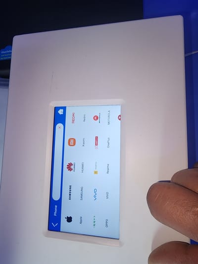 The image shows a close-up of a touchscreen display of a white device, likely a mobile phone or tablet. The screen displays a list of phone brands, including REDMI, Xiaomi, HUAWEI, SAMSUNG, Apple, OPPO, realme, OnePlus, and MOTOROLA. Some brands have their respective logos displayed next to their names. The screen is tilted and slightly out of focus in some areas. A portion of a person's fingers are visible on the right side of the image, appearing to interact with the touchscreen. The device is placed on a white surface, possibly a table or counter, and a blue background is visible on the right edge. The overall scene appears to be indoors, with artificial lighting. There is no indication of the location being Ushtobe, Kazakhstan.