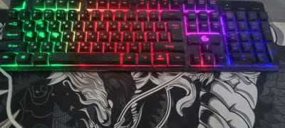 This is a close-up, top-down view of a gaming keyboard with RGB backlighting. The keyboard is dark with black keys, and the lighting shifts through green, yellow, red, pink, and blue. The keys display a mix of Latin and Cyrillic characters. Some keys have visible text, including "Tab", "Caps Lock", "Shift", "Ctrl", "Alt", "Fn", "Enter", "Delete", "Home", "Pgup", "PgDn", "Insert", "End", "Num Lock", "Caps", and "Scroll Lock". The keyboard is placed on a black mousepad with a detailed, stylized image of a creature's head, possibly a dragon or lion, in black and white. The image has a gritty, used appearance, with some dust or debris visible on the keyboard. The lighting suggests it might be a dimly lit room, suitable for gaming. There are no people or specific environmental cues to indicate the time of day, weather, or specific location beyond the presence of a keyboard and mousepad. The overall impression is of a gaming setup.