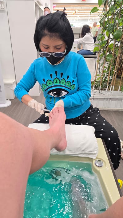 A nail technician, wearing a blue sweatshirt with a large eye graphic and black and white polka dot pants, is performing a pedicure on a client. The technician has black hair, wears glasses and a black face mask, and is using clippers to trim the client's toenails. The client's feet are partially submerged in a bright turquoise pedicure bath filled with bubbly water. A white towel is placed on the edge of the bath. The scene takes place in a brightly lit nail salon, likely in the city of Milton, Canada, indicated by the presence of other patrons and salon equipment visible in the background.