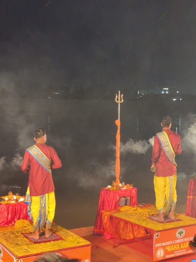The image depicts a religious ceremony taking place at night, likely on the banks of the Tawi River in Jammu, India, given the presence of the "Shree Raghunath Ji Corridor Vikas Council" and "Maha Aarti" text visible on a structure. Two men, dressed in traditional Indian attire consisting of maroon kurtas and yellow dhotis with decorative blue and gold sashes, stand on raised platforms. Both men appear to be participating in a ritualistic offering or prayer, with one holding a small metal vessel. In the center of the scene, a tall trident, or 'trishul', is erected, adorned with a vibrant orange cloth. Smoke or mist is present around the figures and the river in the background, creating a slightly ethereal atmosphere. The platforms are decorated with yellow flowers, and red fabric drapes are visible. The scene is illuminated by artificial lights, highlighting the ceremonial aspects.